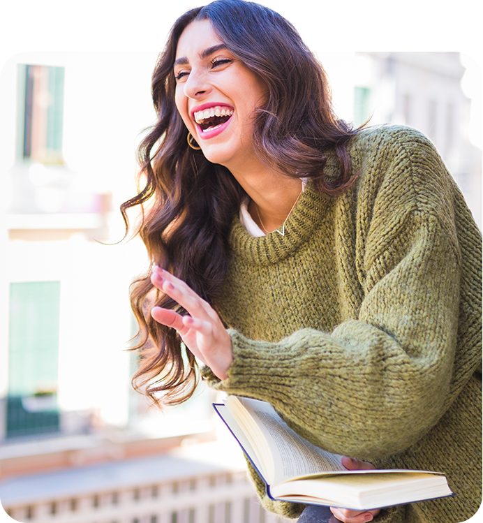 Woman in cosy green jumper laughing while holding an open book; urban background; candid, joyful moment.