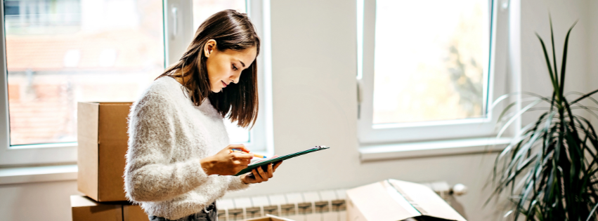 Woman in a cosy jumper, standing in a bright room, checks a clipboard near moving boxes and a large potted plant.