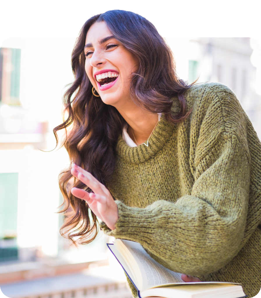 Woman in green jumper laughing while holding an open book, enjoying a sunny day outdoors.