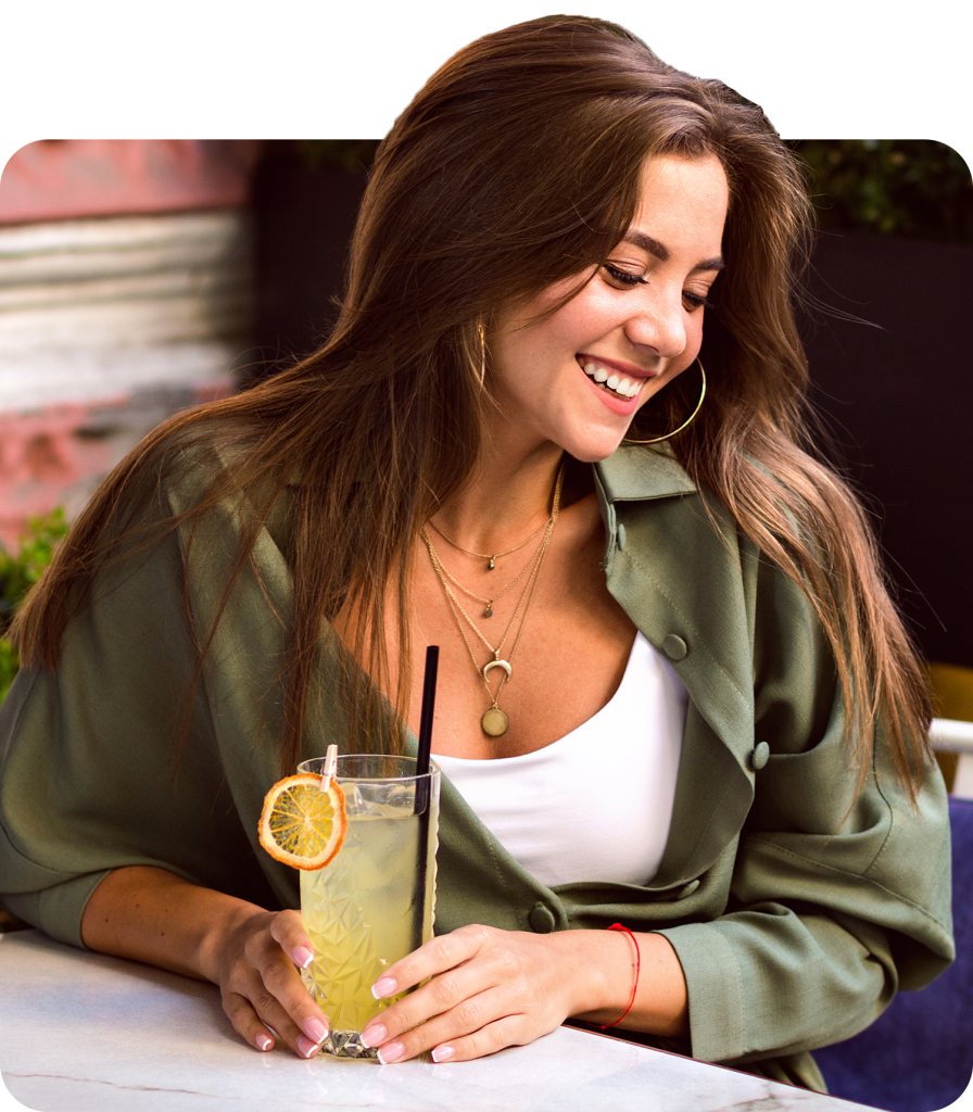 Young woman with a refreshing drink, smiling and enjoying a sunny day outdoors in a green jacket and layered necklaces.