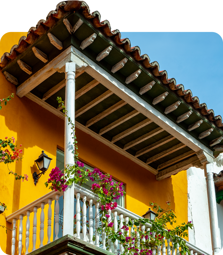 Colourful balcony with blooming flowers and rustic architecture against a bright yellow wall and blue sky.